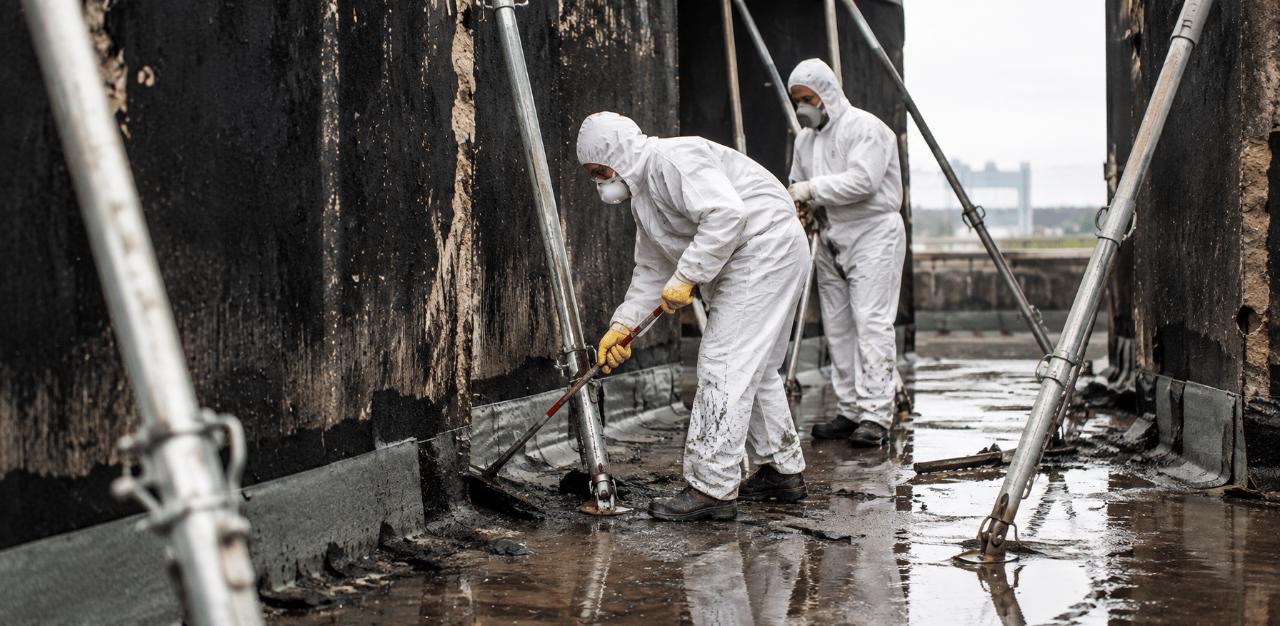 Zwei Mitarbeiter in Schutzanzügen und Atemschutzmasken entfernen Brandrückstände auf einer verrußten, mit Stahlstützen gesicherten Fläche.